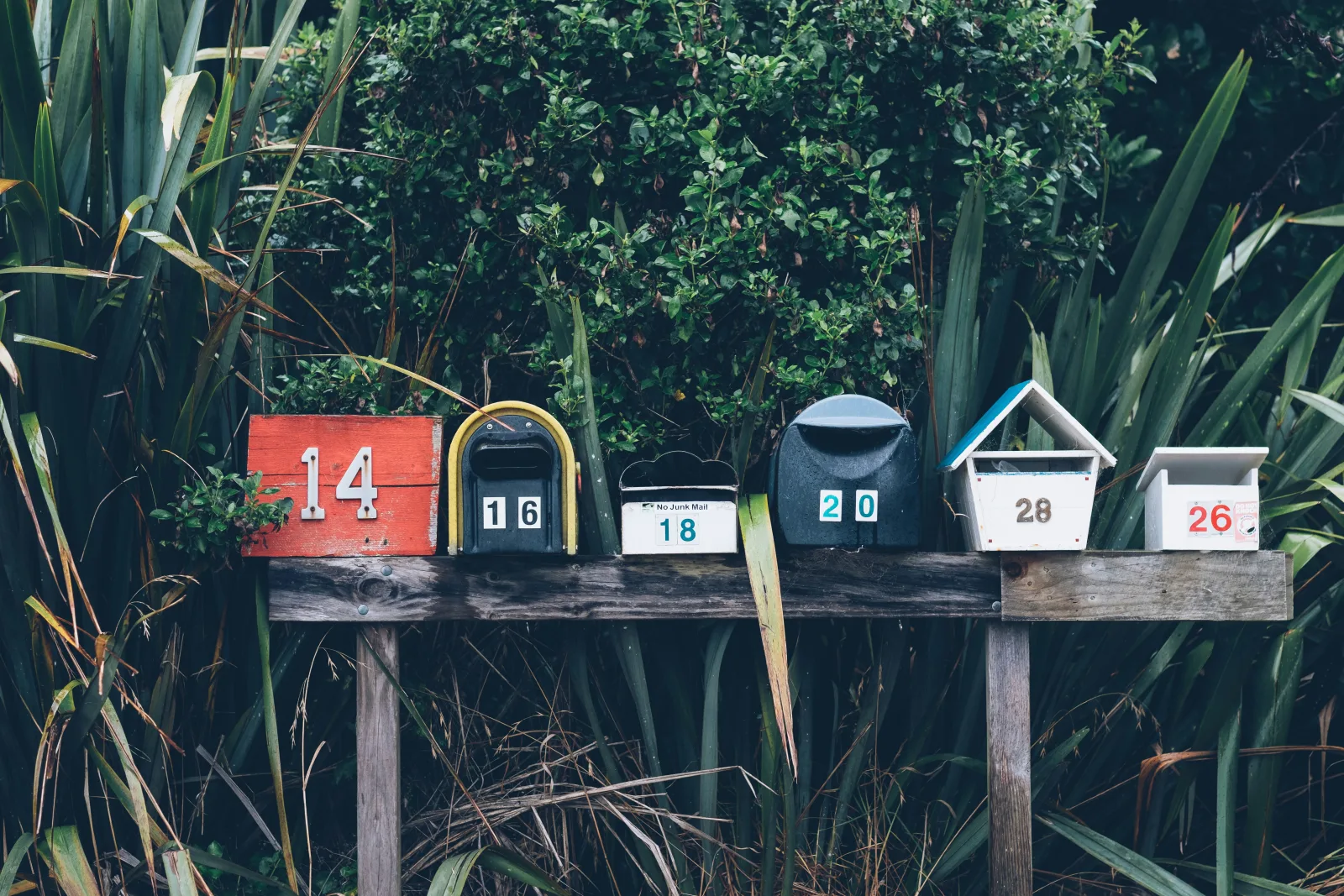 Six mailboxes of different shapes and colours lined up on a wooden rail