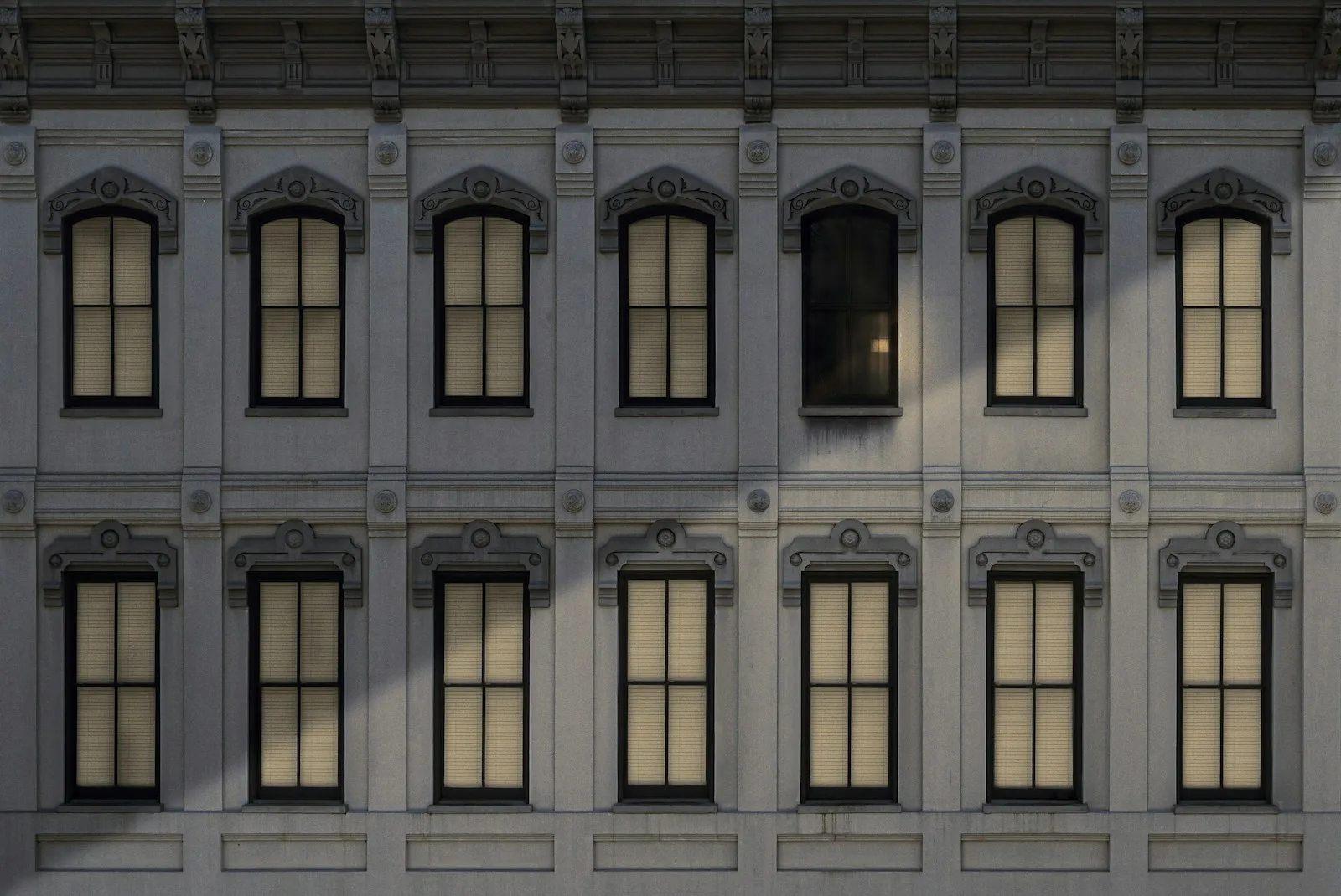 Two rows of identical windows with drawn blinds on a gray facade, with one dark window in the upper row