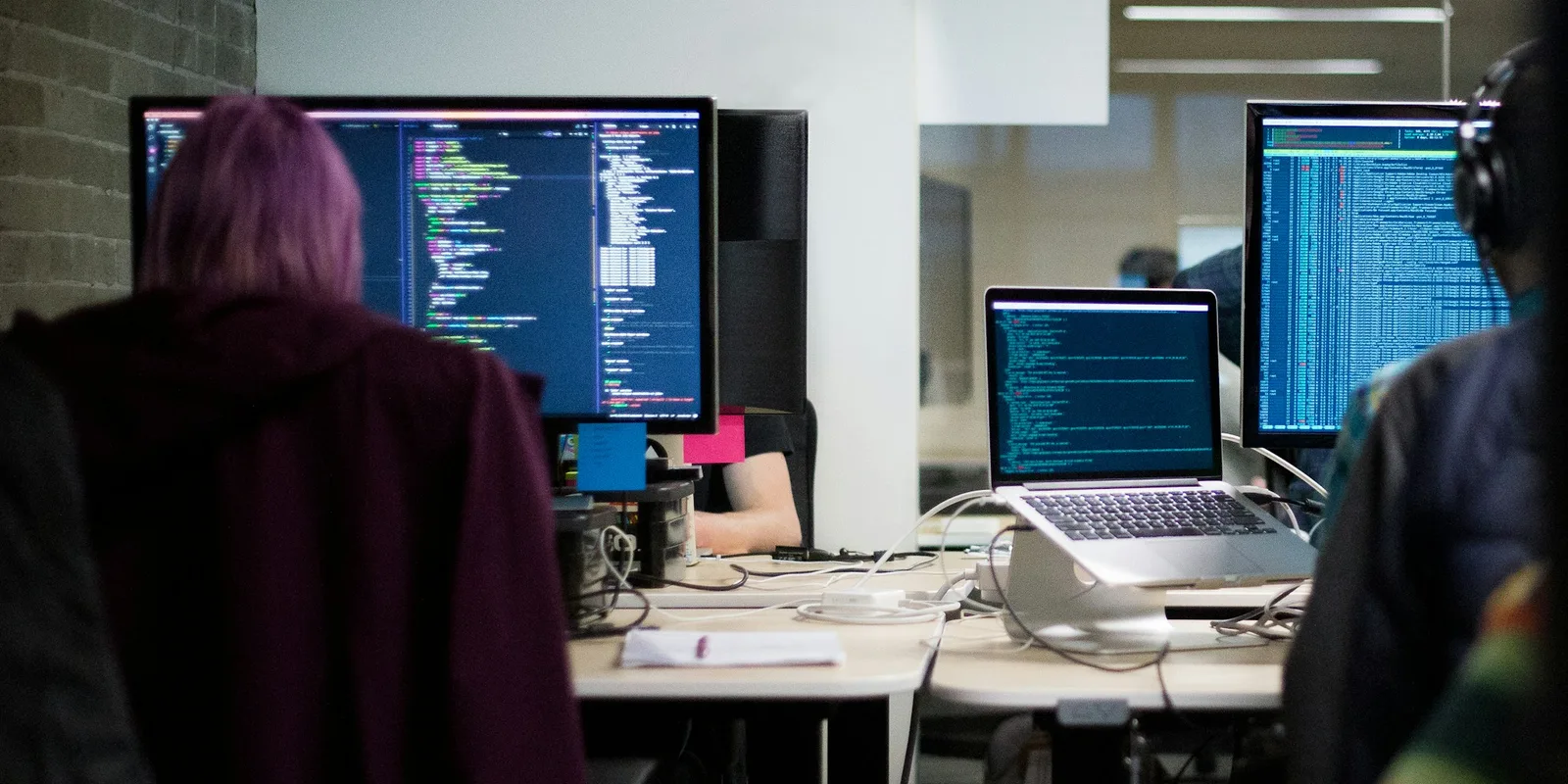 Two developers seen from behind at monitors showing code, with a laptop on a stand, in an open office