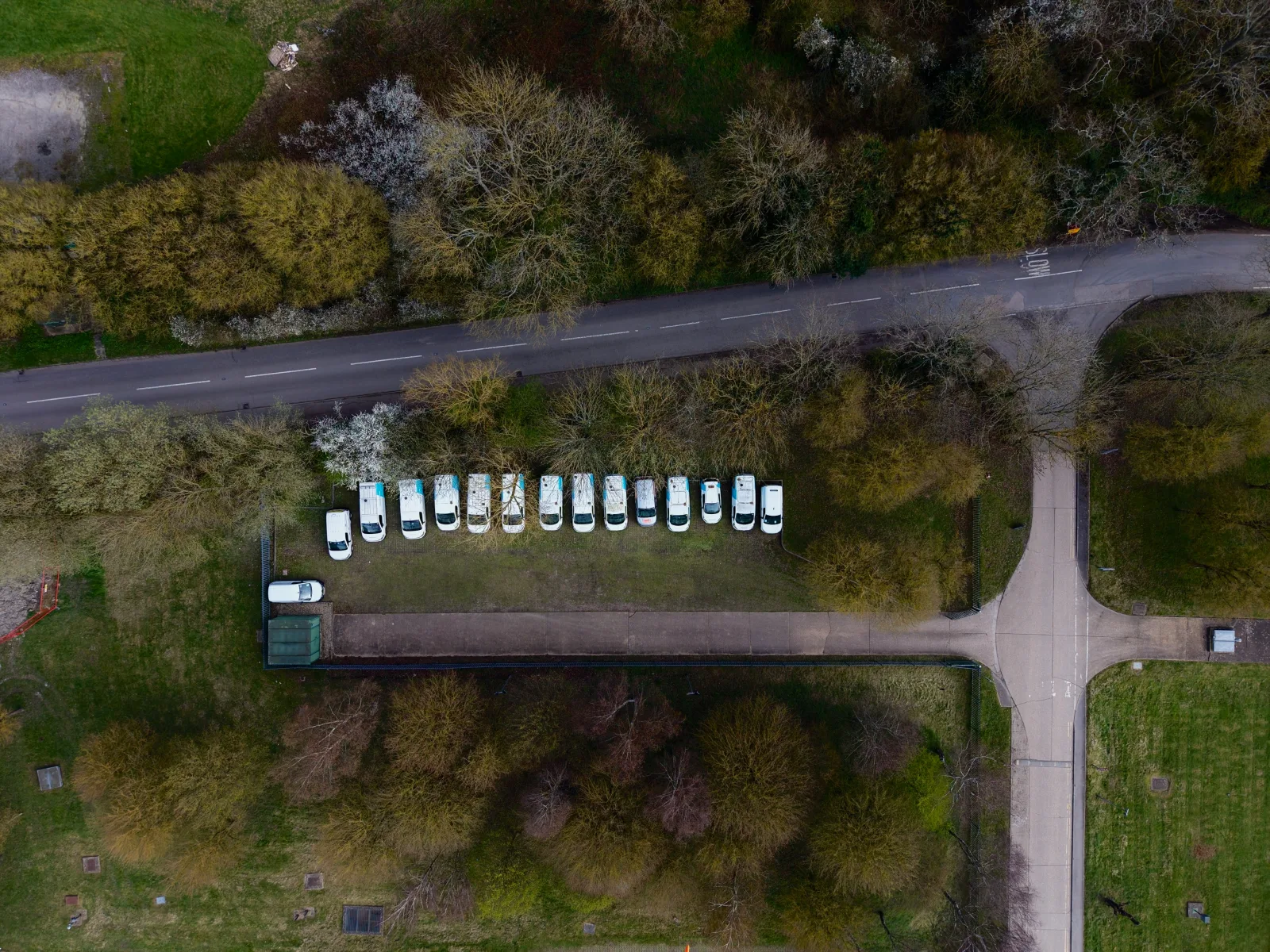 Drone view of a row of white vans parked on grass beside a road