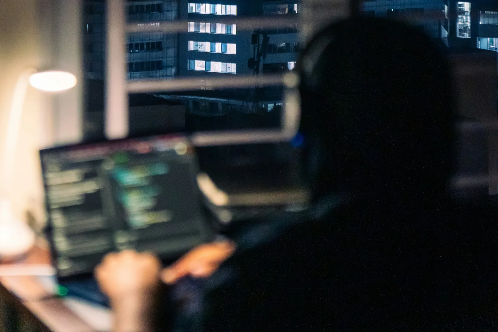 A developer seen from behind wearing headphones, coding on a laptop at night by a window that looks out on lit office buildings