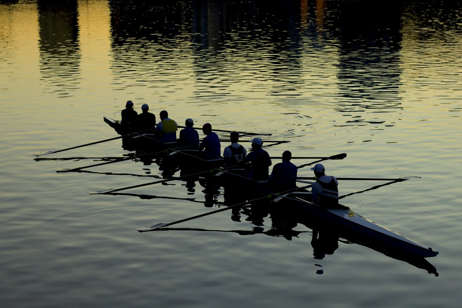 A rowing crew seated in one long boat with their oars resting on still water at dusk